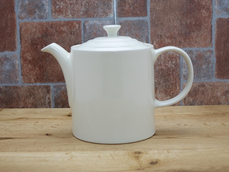 May include: A white ceramic teapot with a lid and handle, set against a brick wall background. The teapot has a spout for pouring and a small knob on the lid. The teapot is sitting on a wooden surface.