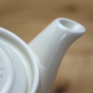 May include: Close-up of a white ceramic teapot, showcasing the spout and lid. The teapot has a glossy finish and a curved spout. The lid has a slightly raised design. The background is a blurred wooden surface.