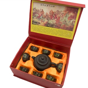 May include: A dark brown clay teapot and seven matching small tea cups with floral patterns, presented in a red gift box. The box lid features a colourful illustration of figures in a landscape and Chinese text, with additional text printed below.