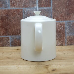 May include: Cream-colored ceramic teapot with a lid and handle. The teapot is cylindrical with a curved handle and a small knob on the lid. The background features a brick wall and a wooden surface.