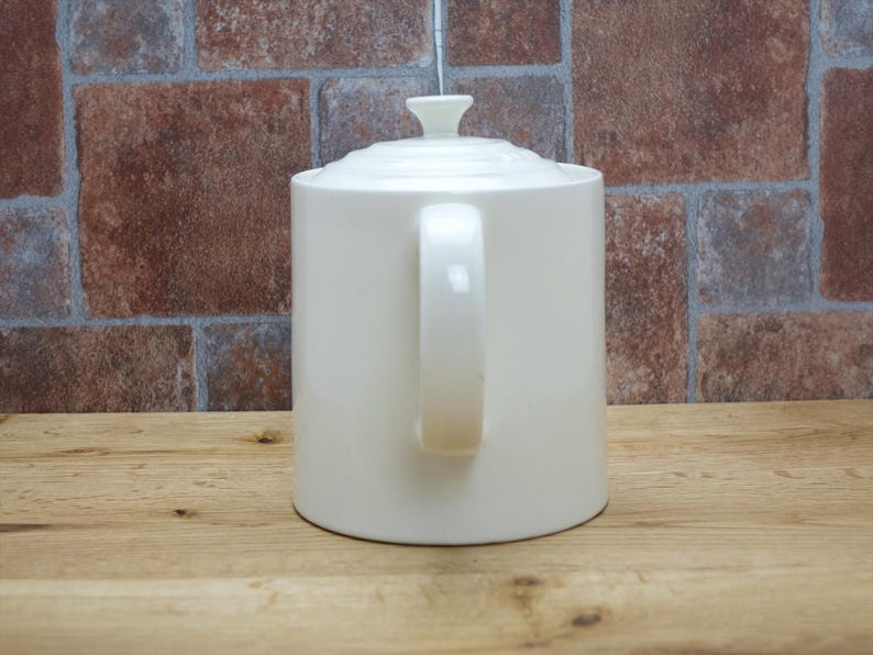 May include: Cream-colored ceramic teapot with a lid and handle. The teapot is cylindrical with a curved handle and a small knob on the lid. The background features a brick wall and a wooden surface.