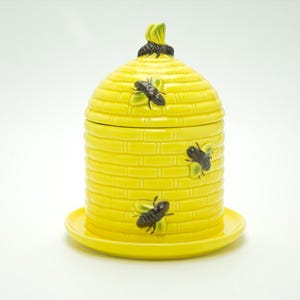 May include: A yellow ceramic beehive-shaped jar with a lid, on a matching saucer. The jar is decorated with three black and yellow bee figurines. The lid has a green and black accent.