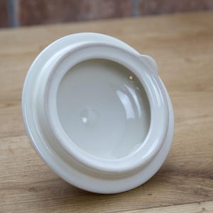 May include: A white ceramic lid with a rounded shape and a small handle. The lid has a glossy finish and a recessed interior. The lid is likely for a teapot or similar kitchenware item.