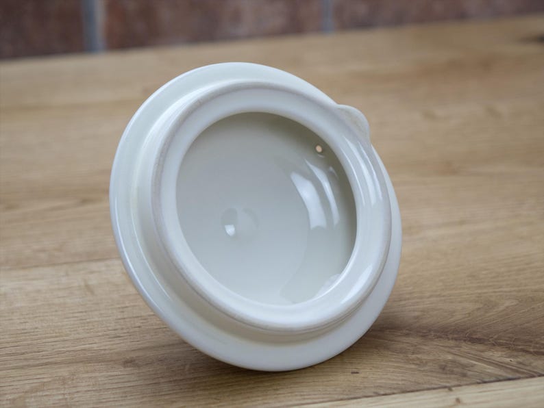 May include: A white ceramic lid with a rounded shape and a small handle. The lid has a glossy finish and a recessed interior. The lid is likely for a teapot or similar kitchenware item.