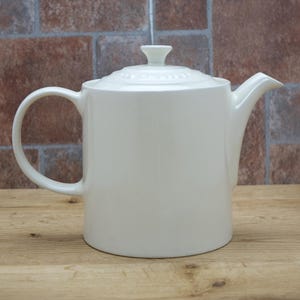 May include: A white ceramic teapot with a lid and handle. The teapot has a curved spout and a rounded body. The background is a brick wall and a wooden surface.