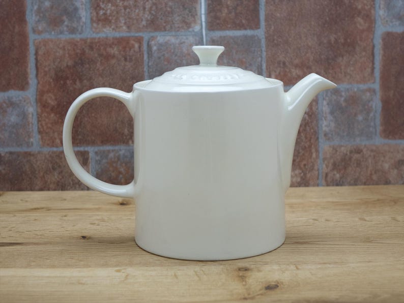 May include: A white ceramic teapot with a lid and handle. The teapot has a curved spout and a rounded body. The background is a brick wall and a wooden surface.