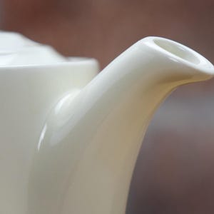 May include: Close-up of a cream-colored ceramic teapot spout. The spout curves gracefully from the body of the teapot, ending in a rounded opening. The teapot has a glossy finish, reflecting the light. The background is blurred, suggesting a focus on the teapot.