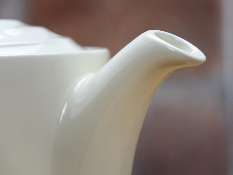 May include: Close-up of a cream-colored ceramic teapot spout. The spout curves gracefully from the body of the teapot, ending in a rounded opening. The teapot has a glossy finish, reflecting the light. The background is blurred, suggesting a focus on the teapot.