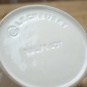 May include: Close-up of the bottom of a white ceramic mug. The embossed text "LE CREUSET" and "MADE IN FRANCE" are visible. The mug has a rounded base and a handle.