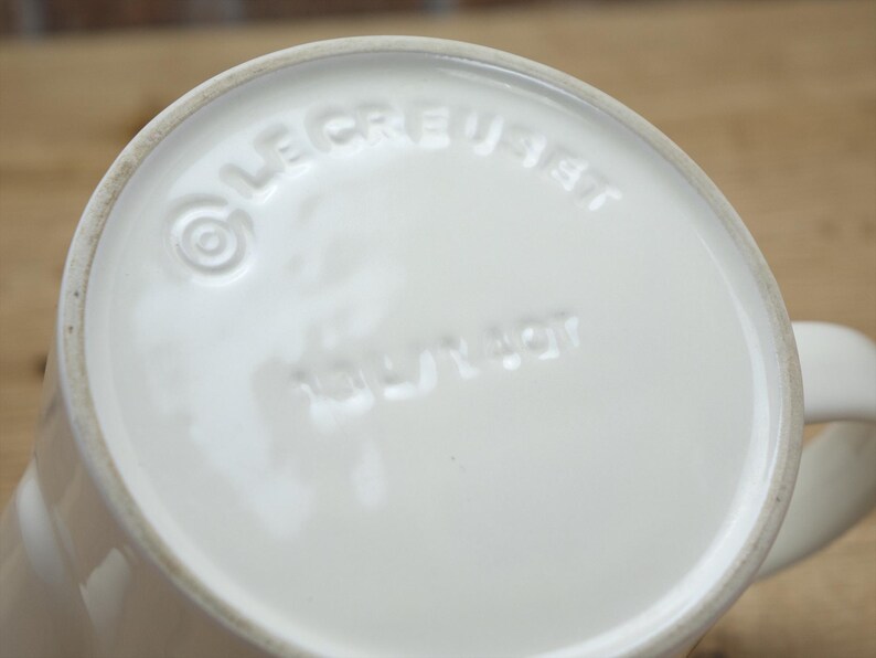 May include: Close-up of the bottom of a white ceramic mug. The embossed text "LE CREUSET" and "MADE IN FRANCE" are visible. The mug has a rounded base and a handle.