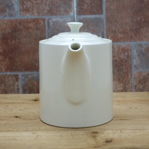May include: Cream-colored ceramic teapot with a lid and spout. The teapot is cylindrical with a rounded base and a small handle on the lid. The background is a brick wall and a wooden surface.
