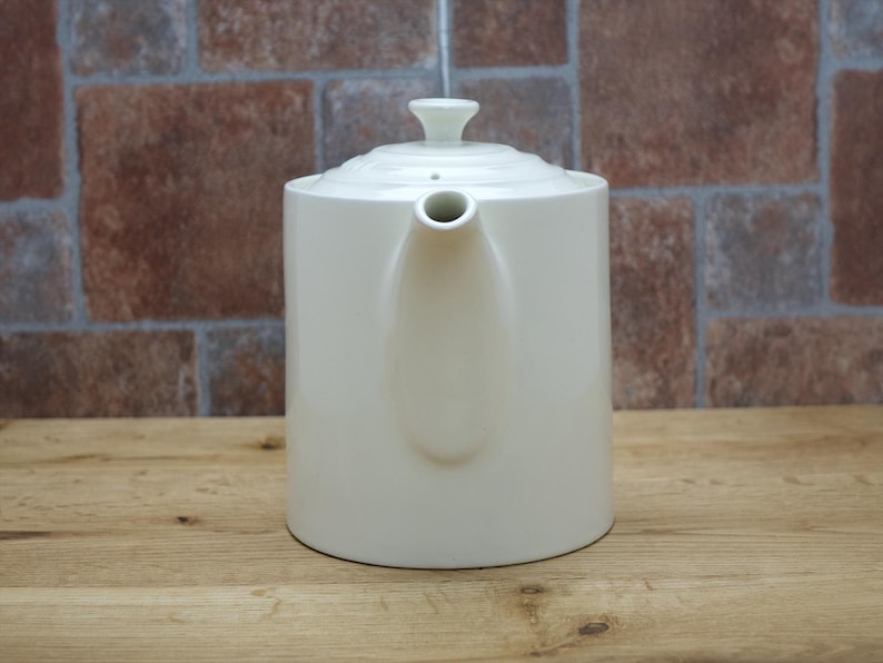 May include: Cream-colored ceramic teapot with a lid and spout. The teapot is cylindrical with a rounded base and a small handle on the lid. The background is a brick wall and a wooden surface.