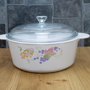 May include: White casserole dish with a clear glass lid. The dish features a floral design with orange, pink, and purple grapes. The dish has two handles and a clear glass knob on the lid. The dish is sitting on a wooden surface.