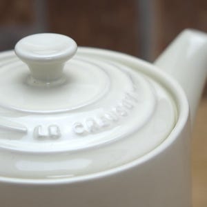 May include: Cream-colored ceramic teapot with a lid and spout. The lid has a small, rounded knob. The brand name "Le Creuset" is embossed on the lid. The teapot is designed with a smooth, glossy finish.