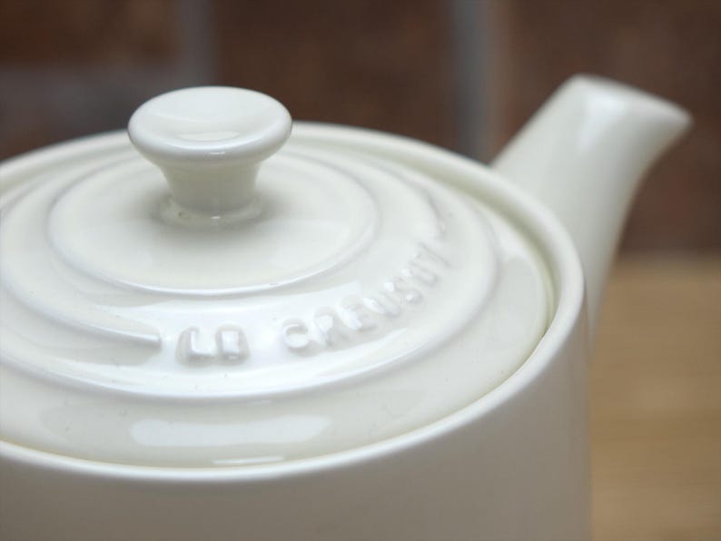 May include: Cream-colored ceramic teapot with a lid and spout. The lid has a small, rounded knob. The brand name "Le Creuset" is embossed on the lid. The teapot is designed with a smooth, glossy finish.