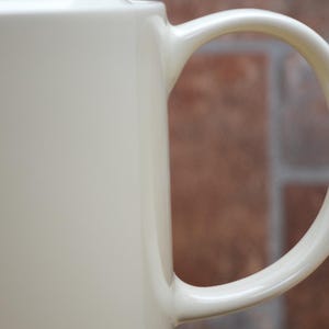 May include: Close-up of a cream-colored ceramic mug with a curved handle. The mug has a smooth, glossy finish. The background is a blurred view of a brick wall.