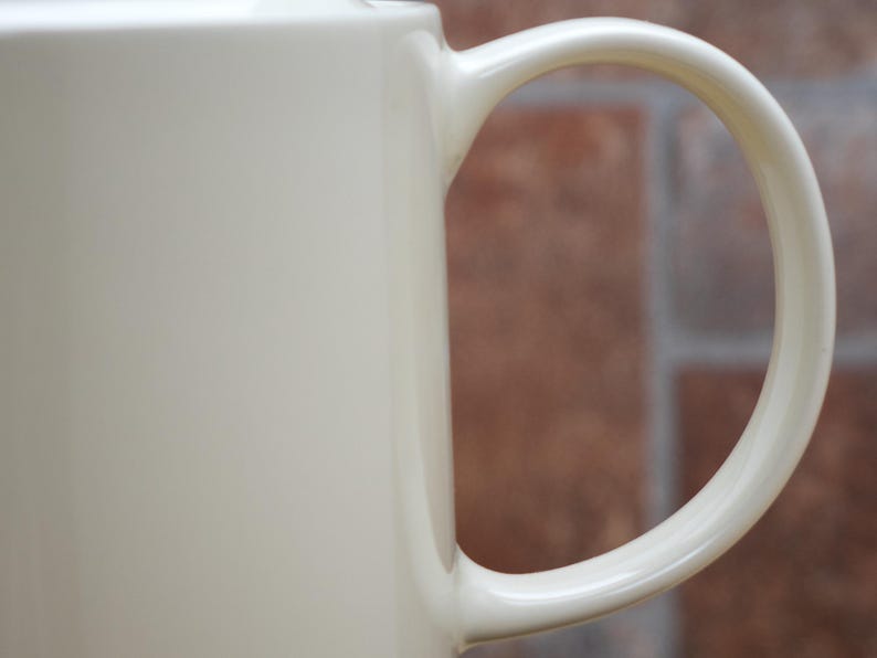 May include: Close-up of a cream-colored ceramic mug with a curved handle. The mug has a smooth, glossy finish. The background is a blurred view of a brick wall.