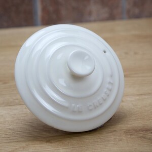 May include: A white Le Creuset lid with a central knob and concentric circular design. The lid has the brand name embossed around the edge. The lid is on a wooden surface.