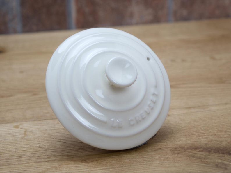 May include: A white Le Creuset lid with a central knob and concentric circular design. The lid has the brand name embossed around the edge. The lid is on a wooden surface.