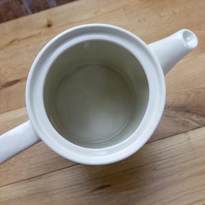 May include: Overhead view of a white ceramic teapot with a handle and spout. The teapot is empty and sits on a wooden surface. The interior of the teapot is a light beige color.