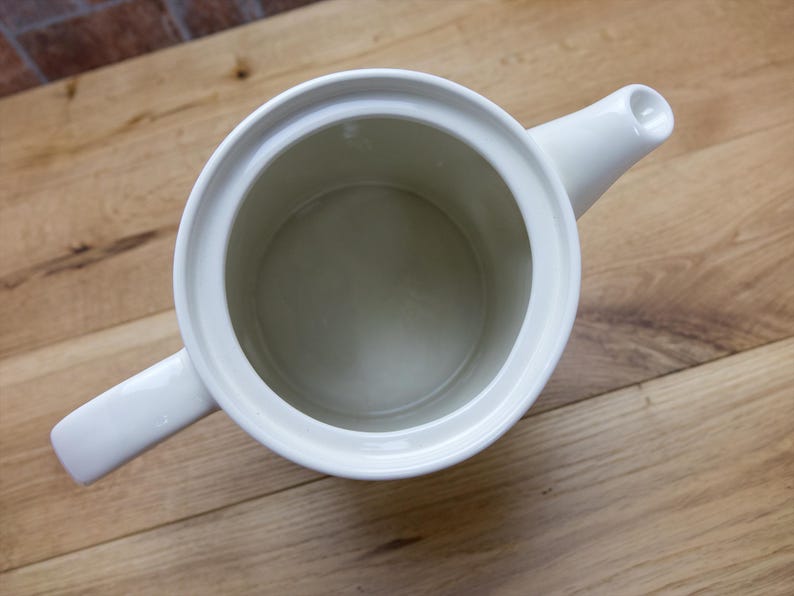May include: Overhead view of a white ceramic teapot with a handle and spout. The teapot is empty and sits on a wooden surface. The interior of the teapot is a light beige color.