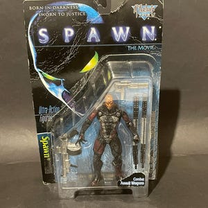 May include: A Spawn The Movie action figure in original packaging. The figure features a dark, textured suit with red and black details. Included accessories are a silver-coloured firearm and two long black combat weapons. The card reads Spawn.