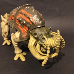 May include: A detailed action figure of a creature with a segmented body, featuring a dark grey carapace with orange markings. The figure has beige limbs and a large, detailed head with blue eyes and sharp teeth. A collectible toy.