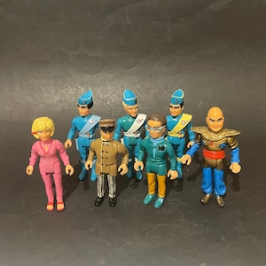 May include: A collection of seven small plastic action figures featuring various characters in distinct outfits. The group includes figures in blue uniforms with caps, a figure in a pink jumpsuit, a tan jacket with striped trousers, and a bald figure in gold armour.