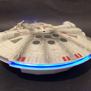 May include: A detailed model of the Millennium Falcon starship, in white and grey with red details. The model has a distinctive circular design and is illuminated with blue lights.