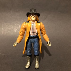 May include: An action figure of a character in a black hat, goggles, and a long tan coat. The figure has a striped shirt, blue trousers, and silver boots. The figure is standing with arms at its sides.
