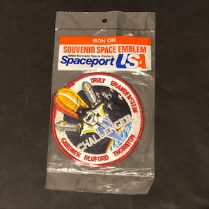 May include: A round, iron-on patch with a blue and white embroidered image of the Space Shuttle Challenger. The patch is surrounded by a red border with the names of the astronauts: Truly, Brandenstein, Gardner, Bluford, and Thornton. The text "Spaceport USA" is printed on the packaging.