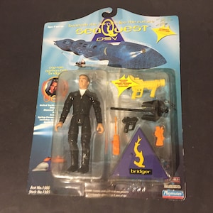 May include: A plastic action figure of Captain Nathan Bridger from the TV show SeaQuest DSV. The figure is wearing a black jumpsuit and is holding a yellow and black gun. The figure is packaged in a plastic blister pack with a blue and yellow background. The packaging includes the SeaQuest DSV logo and the text "Bridger".