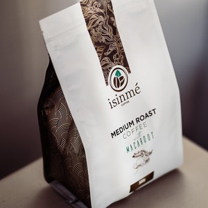 May include: A white bag of coffee with a brown and gold design. The bag is labelled "isinmé Coffee" and "Medium Roast Coffee with Macarroot".