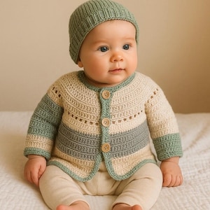 May include: A baby wearing a handmade crochet cardigan and matching beanie. The cardigan features a cream, sage green, and gray color scheme with wooden buttons. The beanie and cuffs of the cardigan are sage green.
