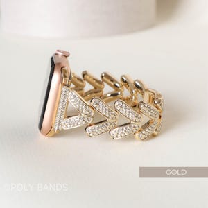 May include: A gold-tone Apple Watch band with a chevron design and clear rhinestones. The band is made of metal and has a polished finish. The band is shown on a white surface. The text "GOLD" is visible in the lower right corner of the image.