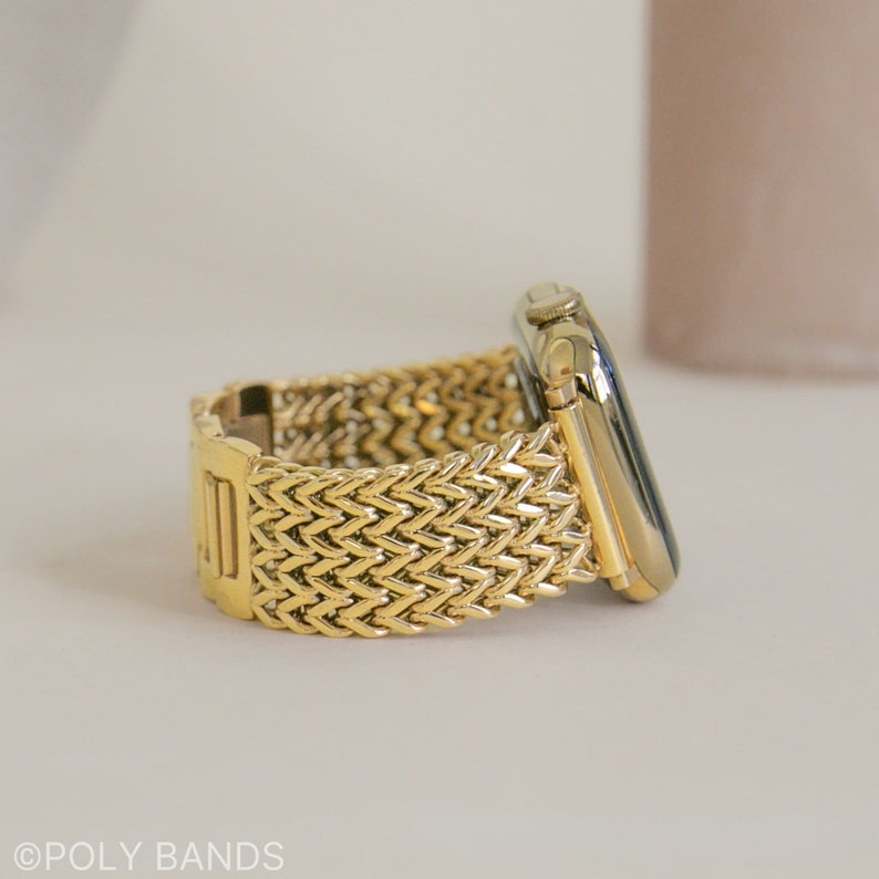 May include: A gold metal Apple Watch band with a woven herringbone pattern. The band is shown on a white surface.