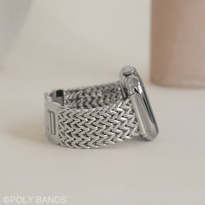 May include: A silver metal watch band with a woven design. The band is attached to a silver Apple Watch.