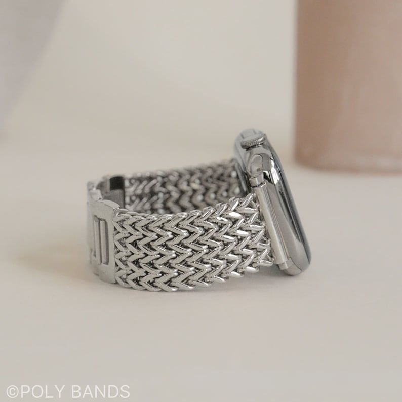 May include: A silver metal watch band with a woven design. The band is attached to a silver Apple Watch.