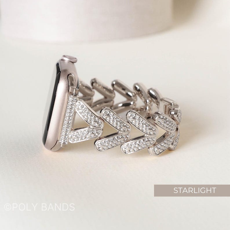 May include: Silver watch band with a clear crystal arrow design. The band is made of metal and has a polished finish. The band is adorned with small, clear crystals that create a sparkling effect. The band is designed to fit a smartwatch.