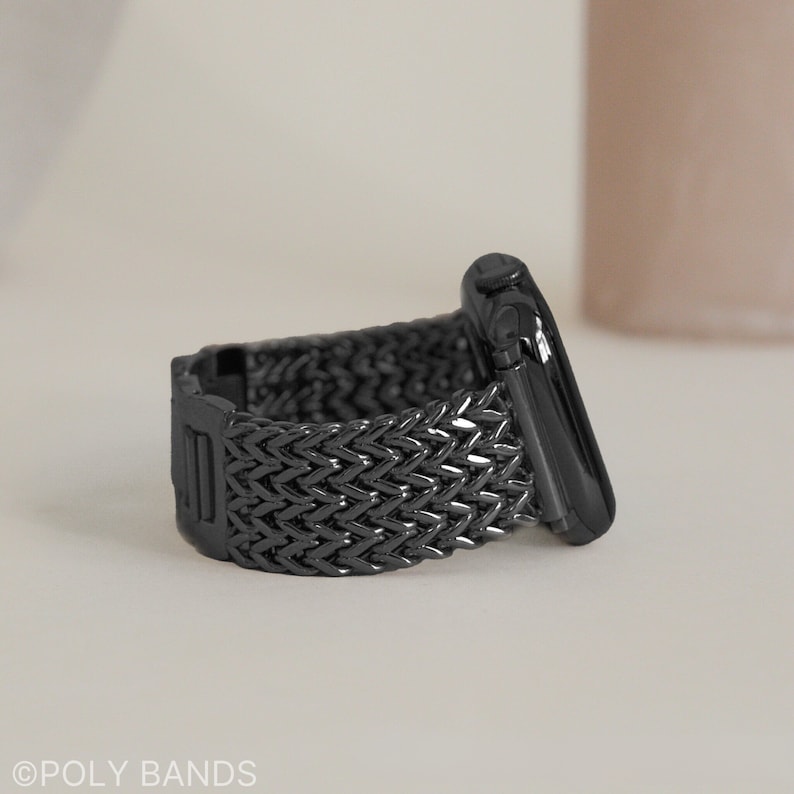 May include: A black braided metal Apple Watch band with a black metal clasp. The band is made of a woven metal design.
