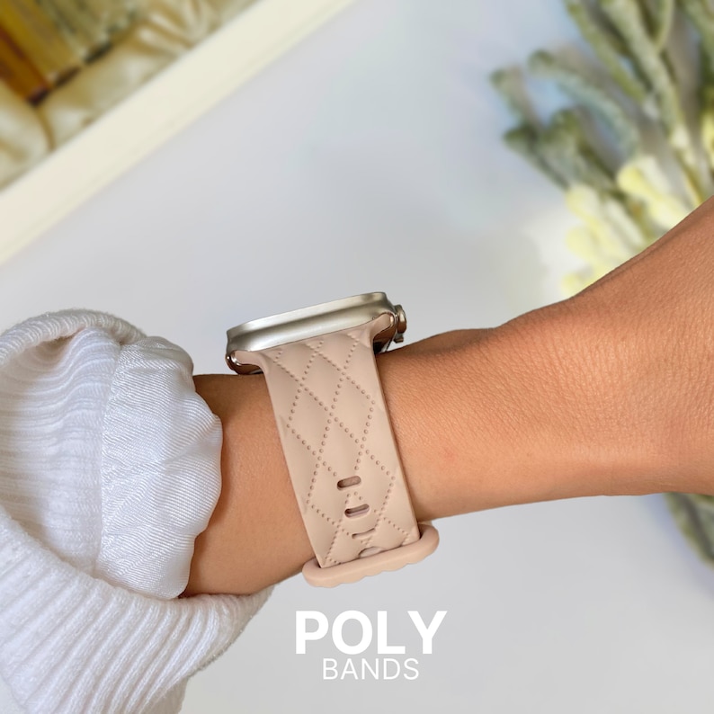 Puede incluir: Una banda de reloj Apple acolchada de color rosa claro con una hebilla plateada. La banda est&aacute; en la mu&ntilde;eca de una persona. El texto "POLY BANDS" est&aacute; en la parte inferior de la imagen.