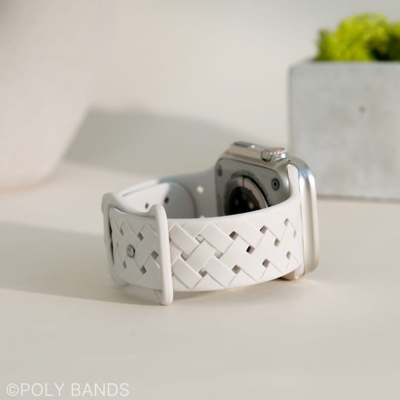 May include: White woven Apple Watch strap with a silver watch face. The strap has a braided design with small holes throughout.