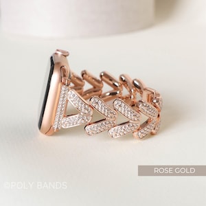 May include: Rose gold Apple Watch band with a diamond-like crystal design. The band is made of metal and has a chevron pattern.