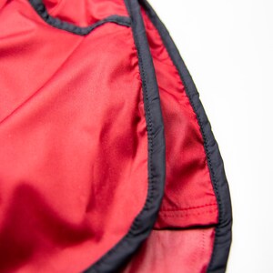 K&ouml;nnte beinhalten: Nahaufnahme von roten Sporthosen mit schwarzem Besatz. Die Shorts bestehen aus einem gl&auml;nzenden, leichten Material und haben einen Kordelzug in der Taille. Die Shorts sind f&uuml;r das Laufen oder andere sportliche Aktivit&auml;ten konzipiert.
