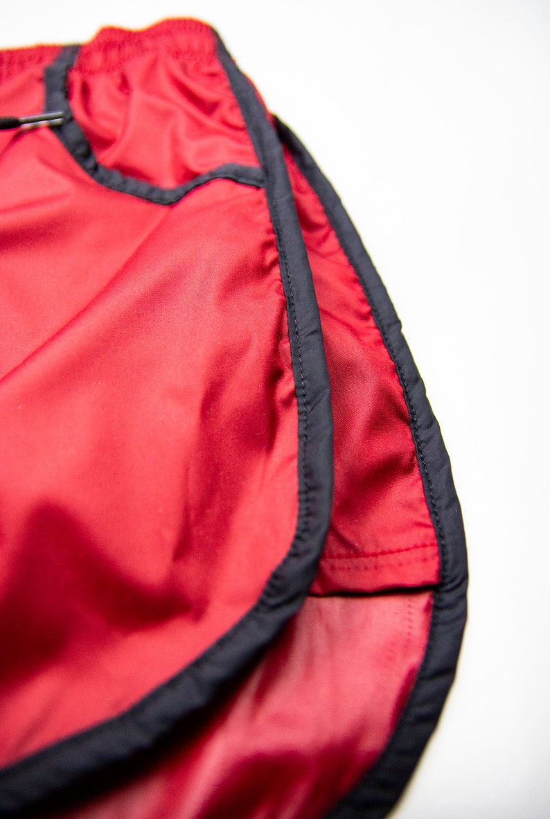 K&ouml;nnte beinhalten: Nahaufnahme von roten Sporthosen mit schwarzem Besatz. Die Shorts bestehen aus einem gl&auml;nzenden, leichten Material und haben einen Kordelzug in der Taille. Die Shorts sind f&uuml;r das Laufen oder andere sportliche Aktivit&auml;ten konzipiert.