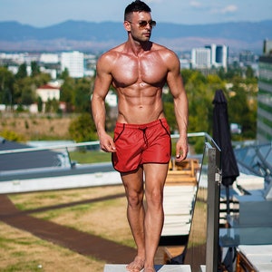K&ouml;nnte beinhalten: Eine muskul&ouml;se Person in roten Badeshorts mit schwarzem Besatz und Sonnenbrille steht in der N&auml;he eines Pools. Die Person befindet sich an einem sonnigen Tag im Freien mit einer Stadt und Bergen im Hintergrund.