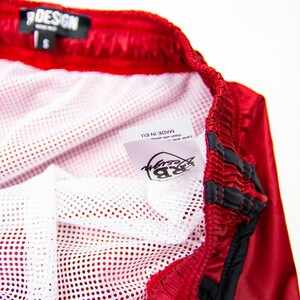 K&ouml;nnte beinhalten: Rote Sporthose mit wei&szlig;em Mesh-Futter. Die Shorts haben einen schwarzen Kordelzug und Zierleisten. Ein Etikett in der Hose tr&auml;gt die Aufschrift "RB Design" und "Made in EU". Die Shorts sind mit der Gr&ouml;&szlig;e "S" gekennzeichnet.