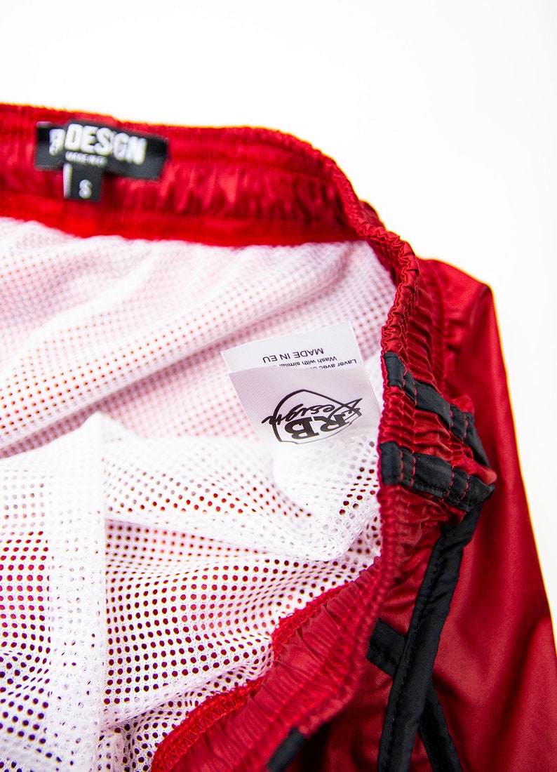 K&ouml;nnte beinhalten: Rote Sporthose mit wei&szlig;em Mesh-Futter. Die Shorts haben einen schwarzen Kordelzug und Zierleisten. Ein Etikett in der Hose tr&auml;gt die Aufschrift "RB Design" und "Made in EU". Die Shorts sind mit der Gr&ouml;&szlig;e "S" gekennzeichnet.