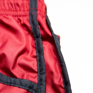K&ouml;nnte beinhalten: Nahaufnahme von roten Sporthosen mit schwarzem Besatz. Die Shorts haben einen Kordelzugbund und eine glatte, leicht gl&auml;nzende Textur. Der schwarze Besatz umrandet die Seiten und N&auml;hte der Shorts.
