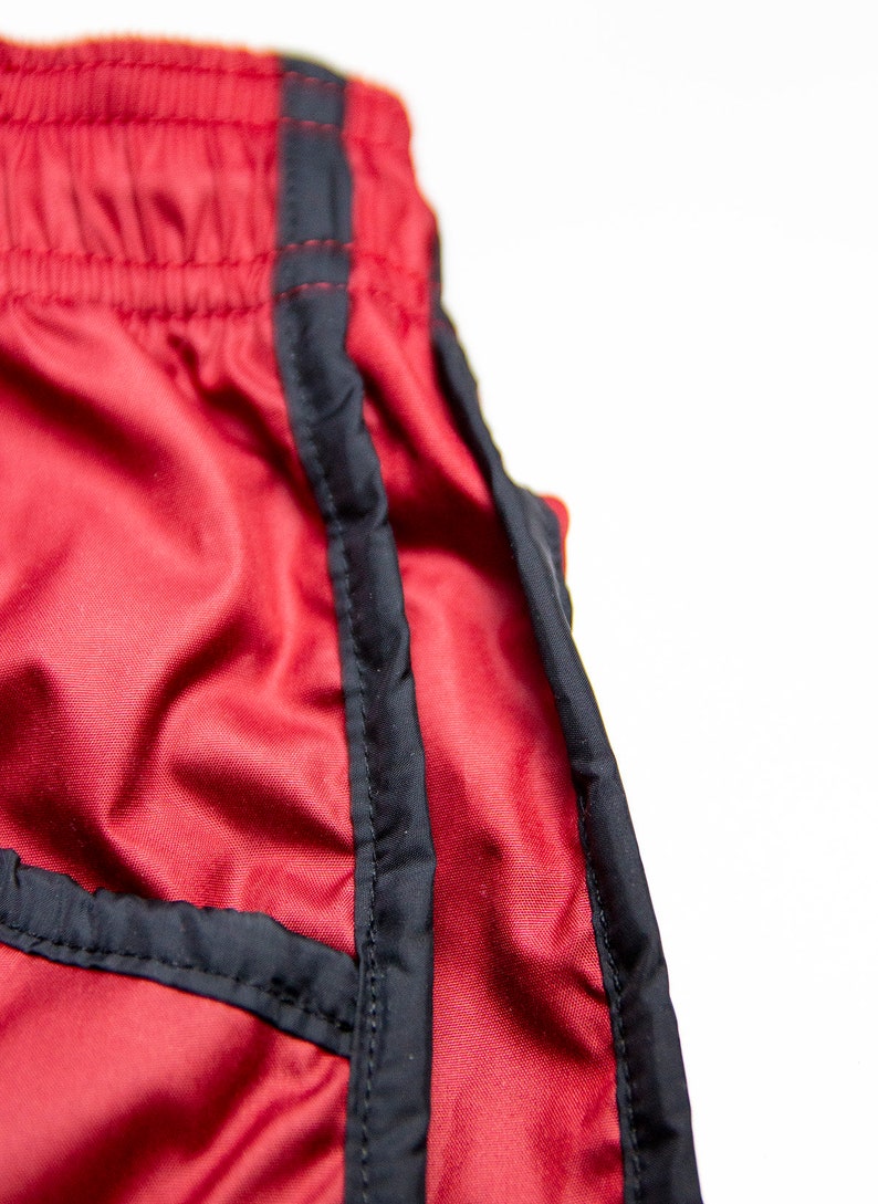 K&ouml;nnte beinhalten: Nahaufnahme von roten Sporthosen mit schwarzem Besatz. Die Shorts haben einen Kordelzugbund und eine glatte, leicht gl&auml;nzende Textur. Der schwarze Besatz umrandet die Seiten und N&auml;hte der Shorts.